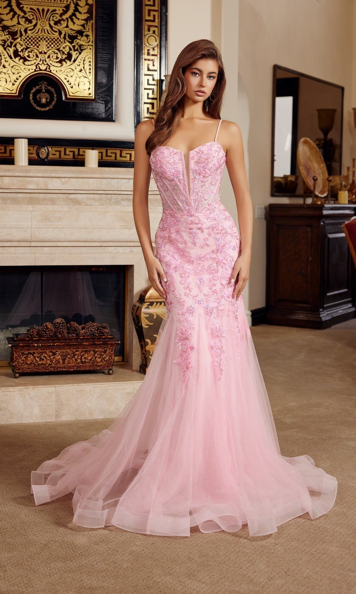 A woman poses elegantly indoors in front of a fireplace, wearing the Nox Anabel H1874 long pink prom dress with beaded sequin embroidery, floral accents, and a tulle skirt.